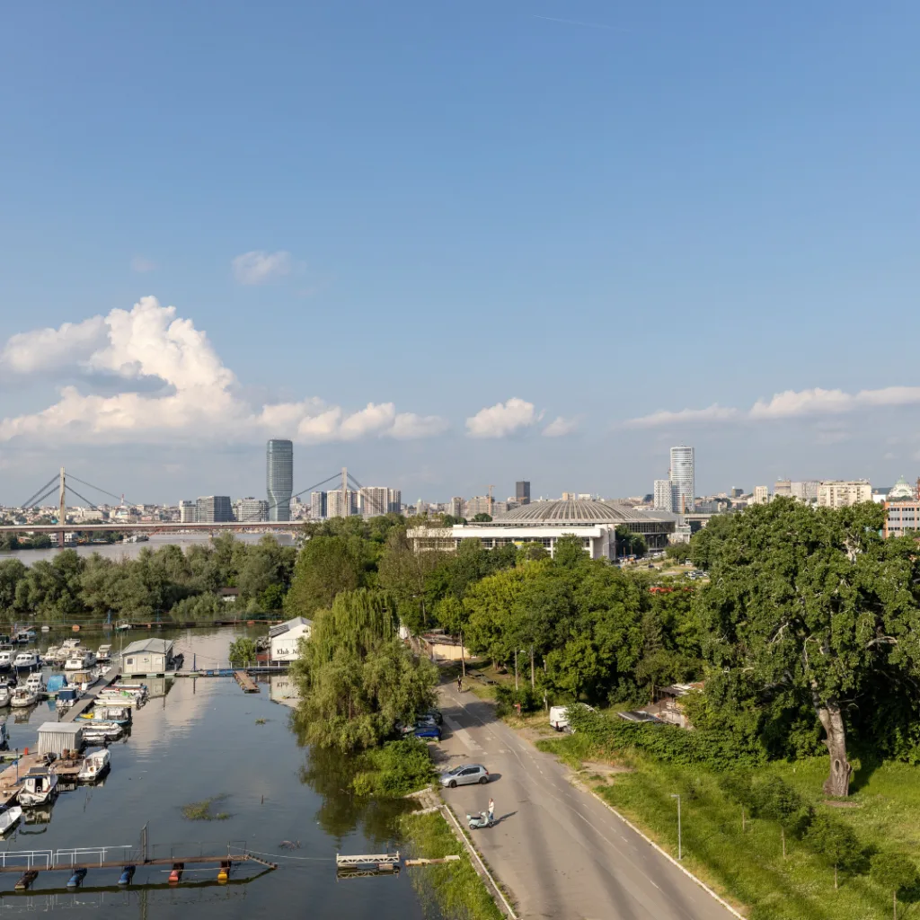 Riverfront cityscape with boats at a marina, a bridge on the left, and a tree-lined road in the foreground under a clear blue sky.